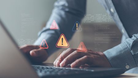 A businessperson's hands using a laptop, with overlaid red and yellow, holographic warning symbols indicating a cyber scam and fraud.