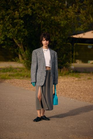 A woman wearing a gray oversized blazer, a white crop T-shirt, gray midi skirt, black shoes, blue leather gloves, and a matching blue top-handle bag.