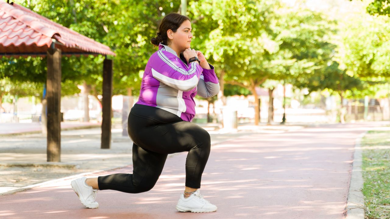 A woman performs a lunging exercise outside. She wears leggings, a long-sleeved sports top and sneakers. Her right foot is in front and her left foot is behind as she assumes the lunge position, with both knees bent. Her hands are clasped underneath her chin. Behind her we see tarmac and a line of trees.