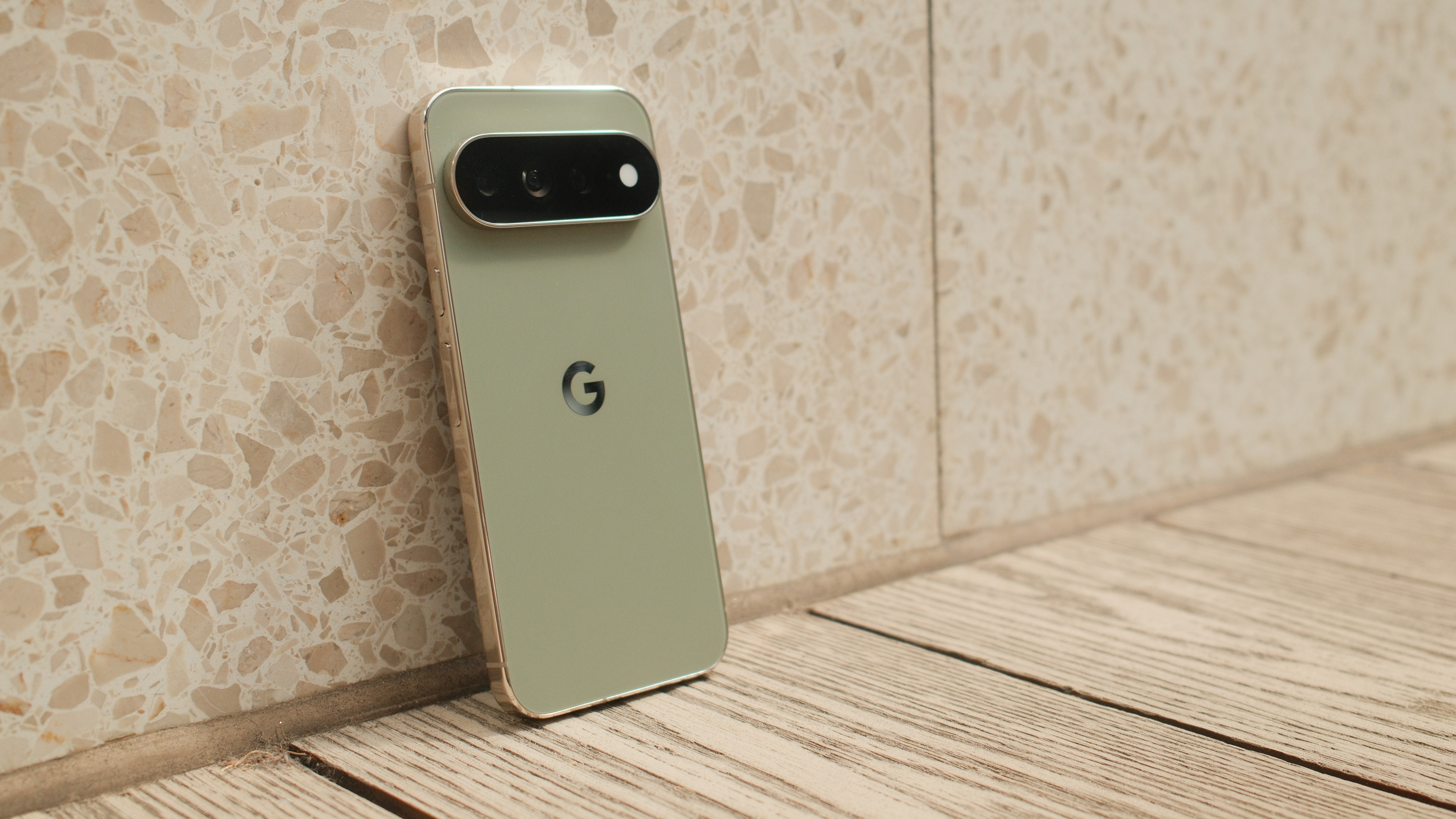 Google Pixel 11 Pro XL leaning against a light stone wall on a wooden floor, showing its rear design and camera bar.