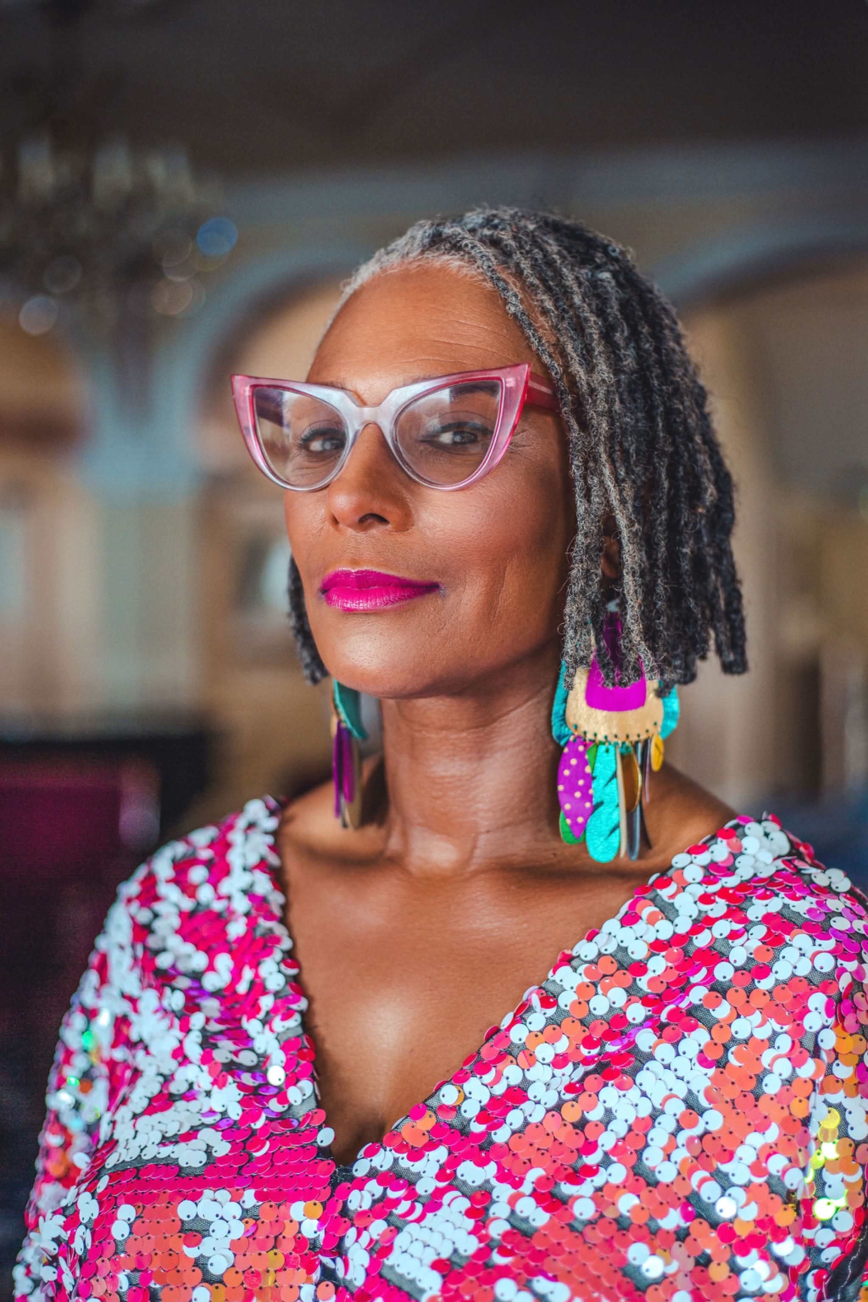 A woman wearing pink glasses, large feathered earrings and a sequined top, looking sideways at the camera