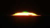 The half dome of a glowing sunset is seen against a black sky with the silhouette of an airplane in the front and a green shape above it. 