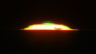 The half dome of a glowing sunset is seen against a black sky with the silhouette of an airplane in the front and a green shape above it. 