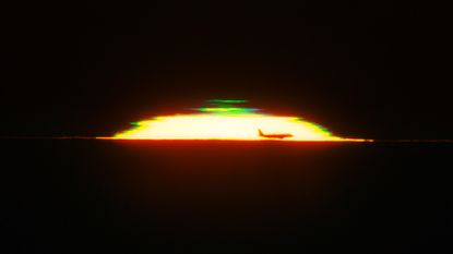The half dome of a glowing sunset is seen against a black sky with the silhouette of an airplane in the front and a green shape above it. 
