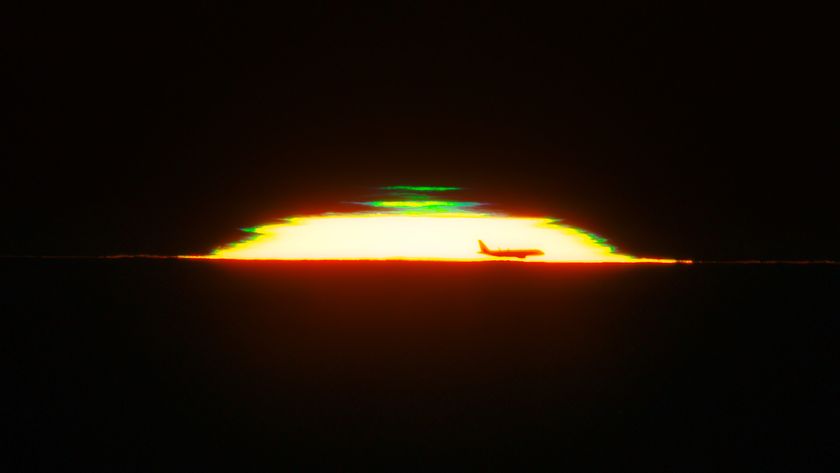 The half dome of a glowing sunset is seen against a black sky with the silhouette of an airplane in the front and a green shape above it. 