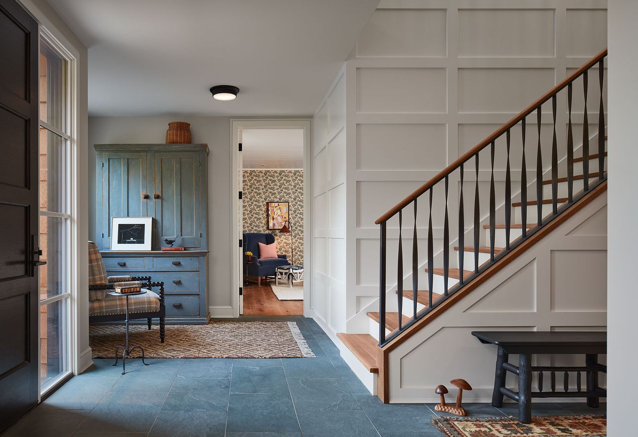 Entrance hall and stairs with white paneled walls, slate tiled floor and blue chest of drawers and armchair in the distance