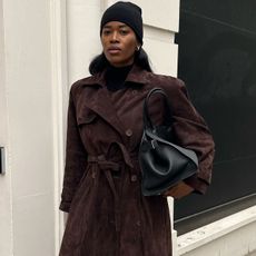 Woman wearing brown suede trench coat, black beanie, and black leather bag leaning against door.