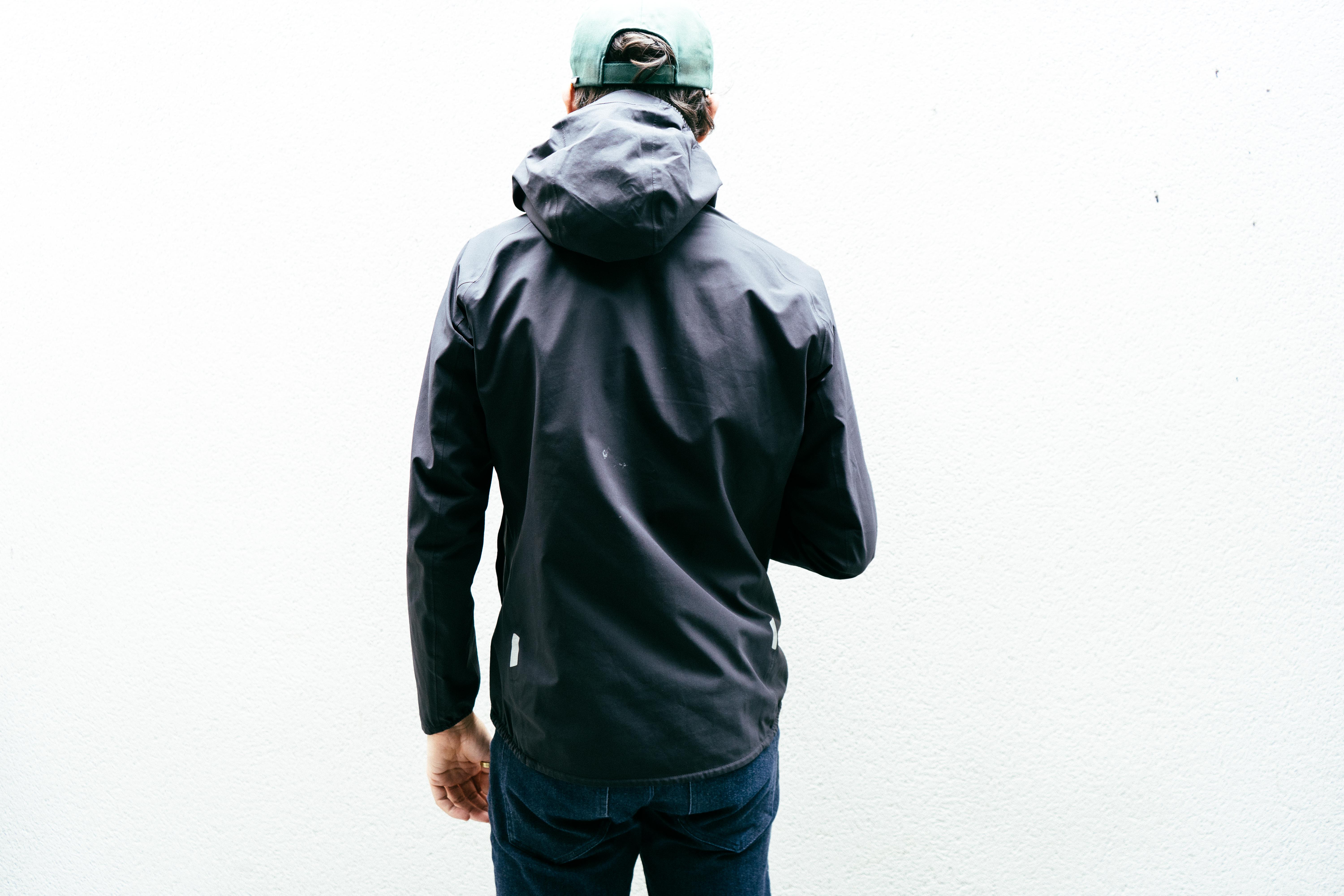 A man in a black waterproof jacket stands facing a white wall