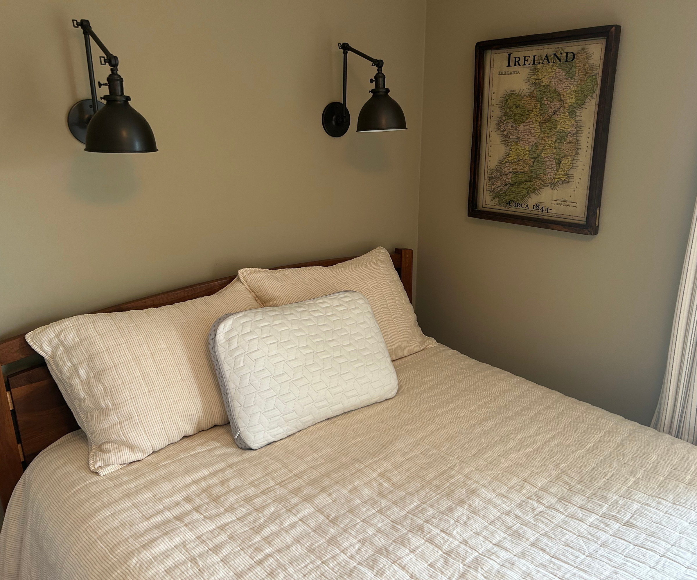 a bed with three pillows, two wall sconces on the wall, and a map in a picture frame