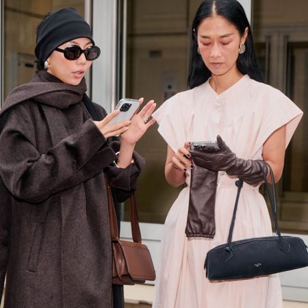 a close street style photo of two women standing outside on the sidewalk scrolling their phones in search of the best ssense sale finds 