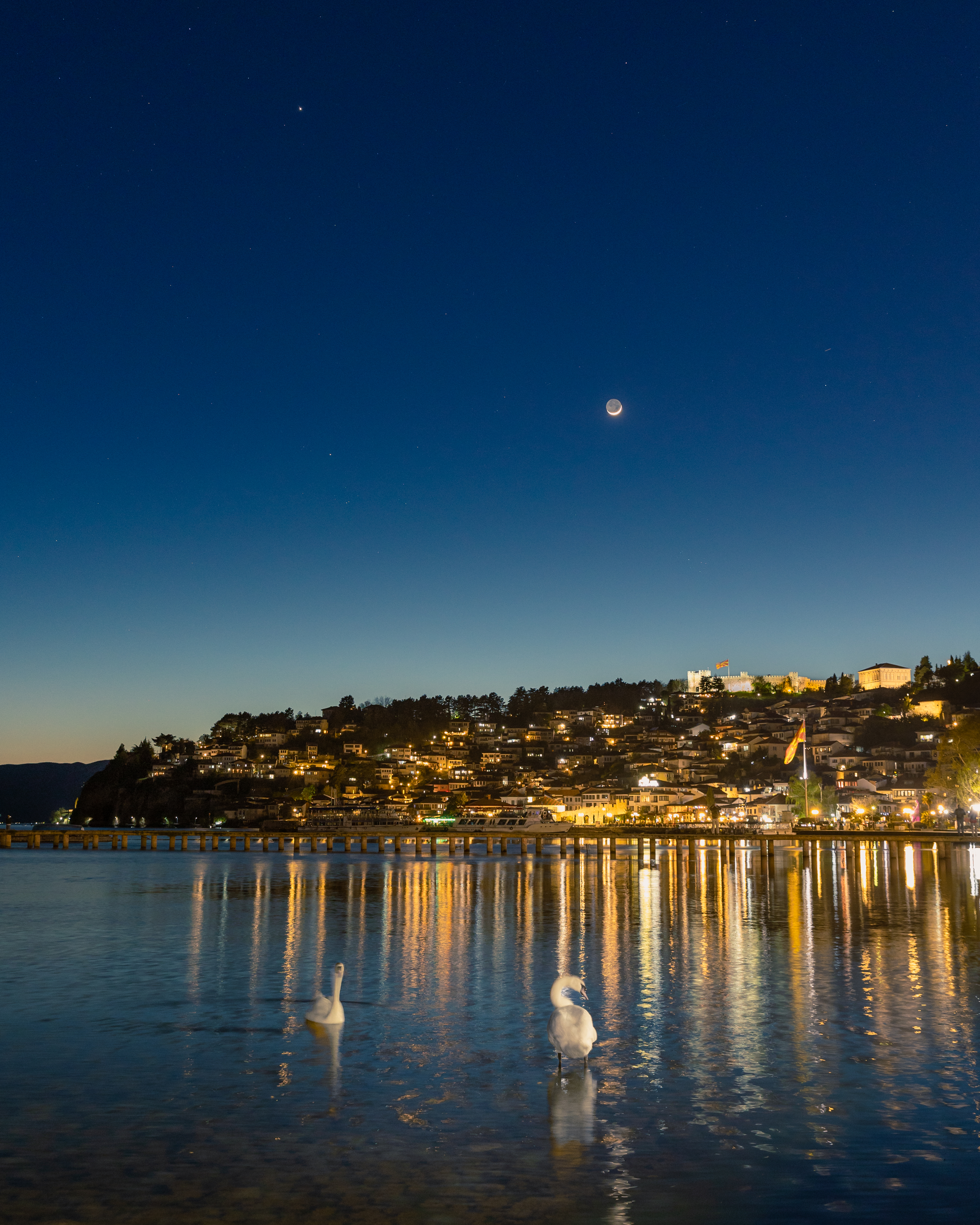 two bright orbs in a deep blue sky above the lights of a small town on a hillside, the lights from which are reflected in a lake