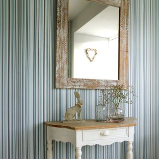 A hallway with a wall covered in a striped wallpaper with a small console table and a wall mirror