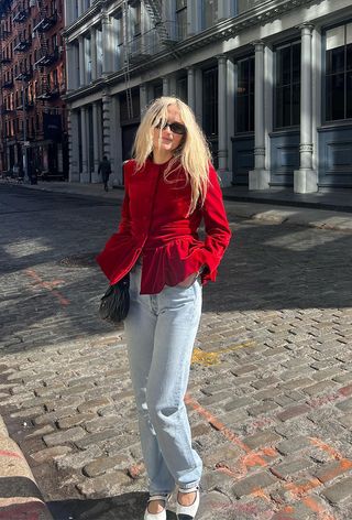 how to wear velvet is shown in a photo of a woman crossing the street in New York wearing black sunglasses with a red velvet blazer, light wash straight-leg jeans, white cap-toe Mary Jane flats, and a black Miu Miu bag