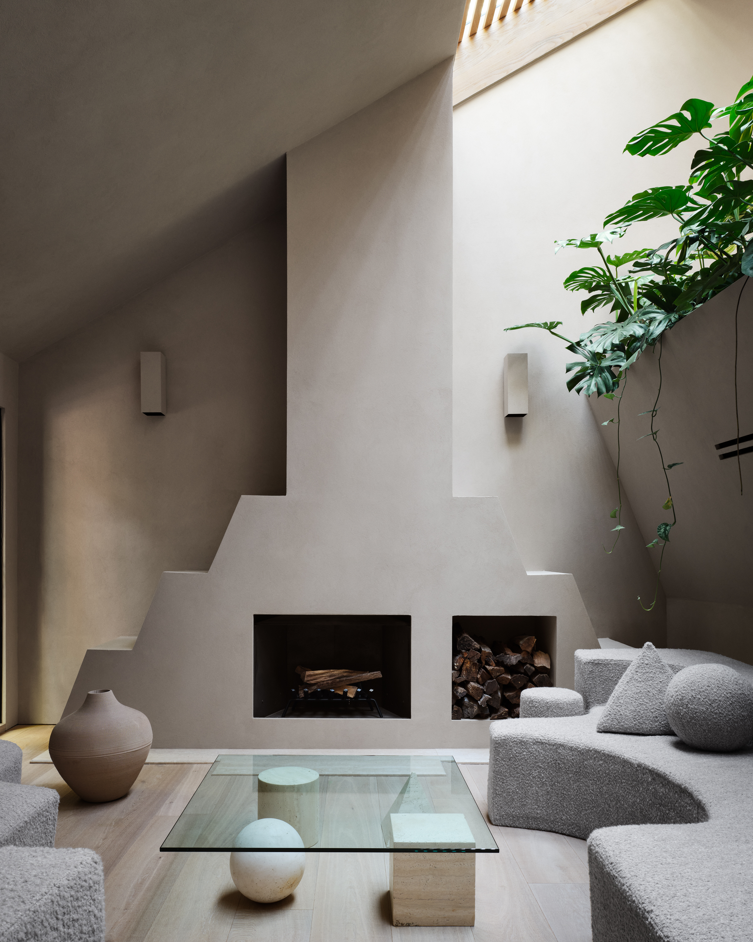 neutral living room with angled ceiling and natural light pouring in, leafy plants spilling down the wall, a gray sculptural sofa, a built-in fireplace with log stacks, a glass coffee table with sculptural stone base, a large pot, and light timber floorboards