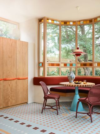 Breakfast nook surrounded by tall, geometric windows with coloured glass inserts. A custom rust coloured bench sits in the alcove of the nook with a wooden table and two rust rattan chairs, with light wooden custom joinery to the left of the seating area.