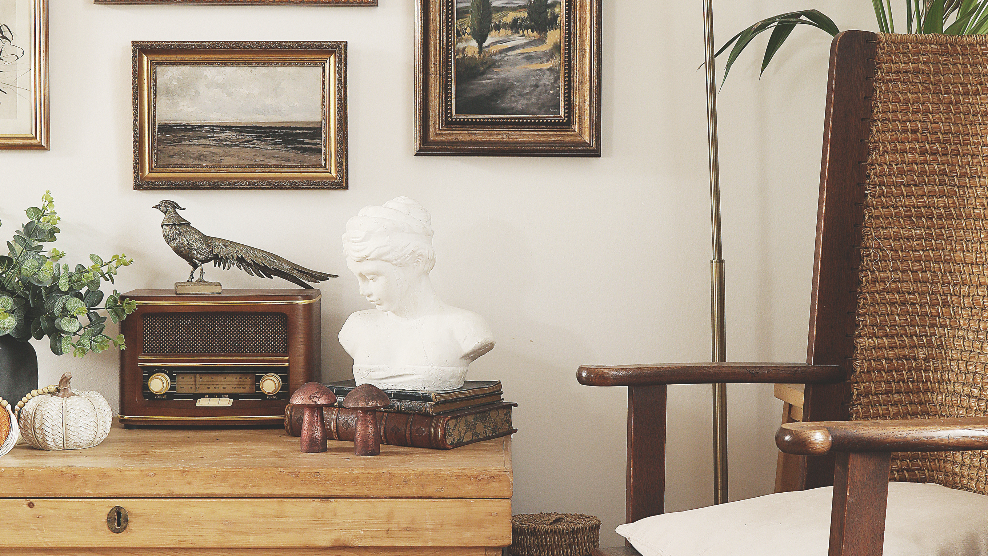 Interior view of a sitting room with a wicker chair and wooden floor and chest with lots of photo frames, retro radio and white bust of a woman.