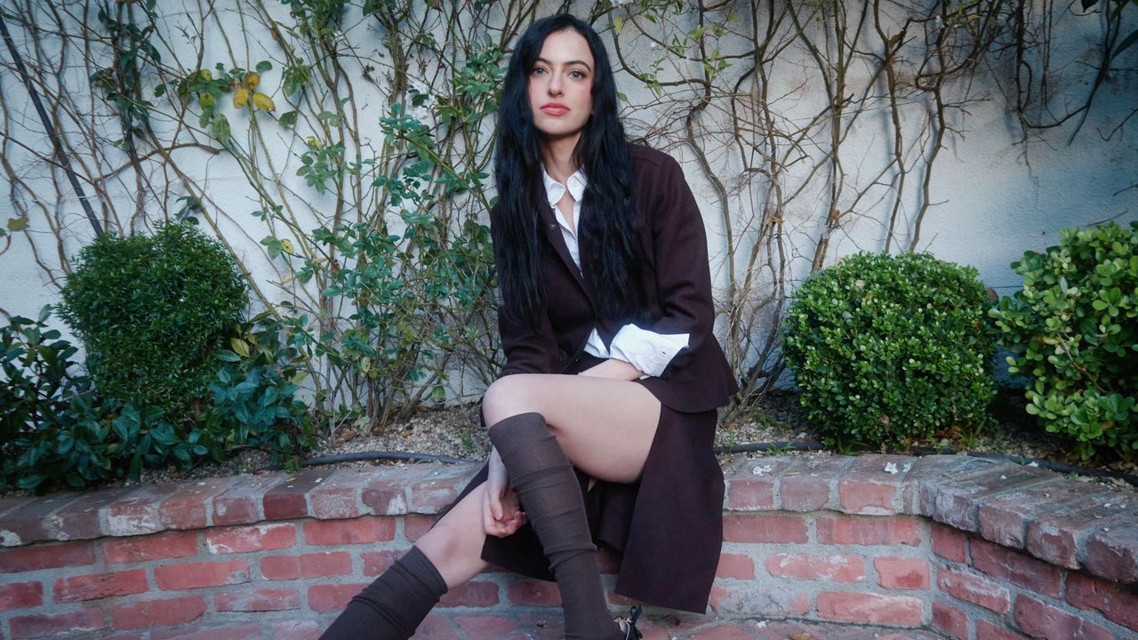 actress/writer cazzie david sits on a brick bench wearing a black blazzer and tall knee socks