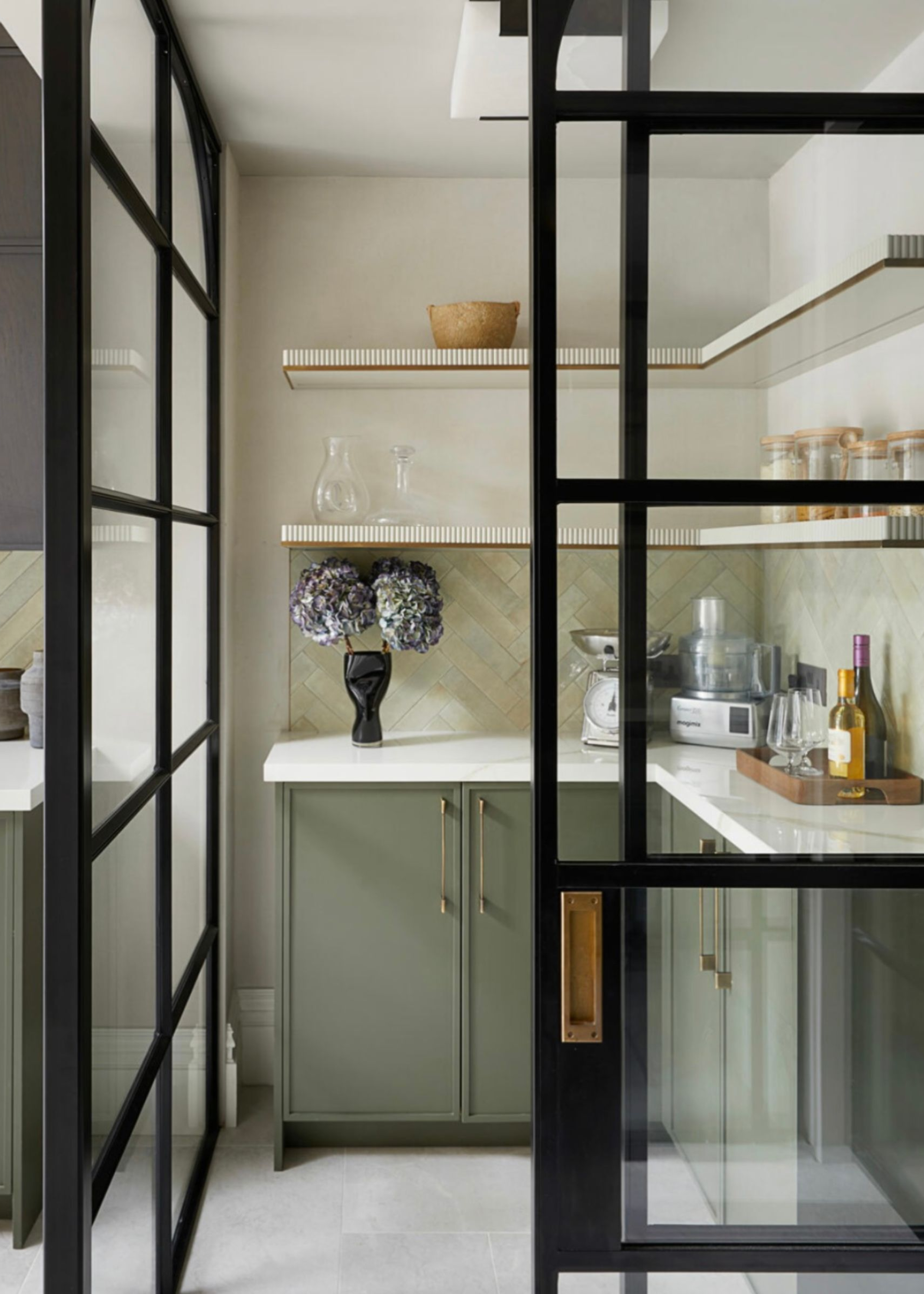 Pantry with Crittall doors, green cabinets, countertop and open shelving. On the shelf is a vase with flowers inside as well as a tray with oil and other kitchen essentials