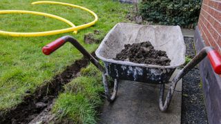 trench dug in garden with yellow pipe nearby and wheelbarrow containing soil