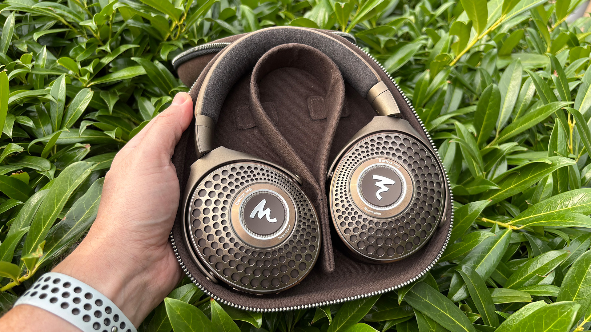 Focal Bathys MG wireless headphones in case held in hand in front of green bush