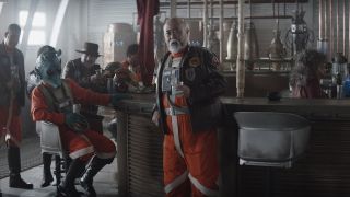 characters in futuristic silver armor talk to one another in a dingy bar