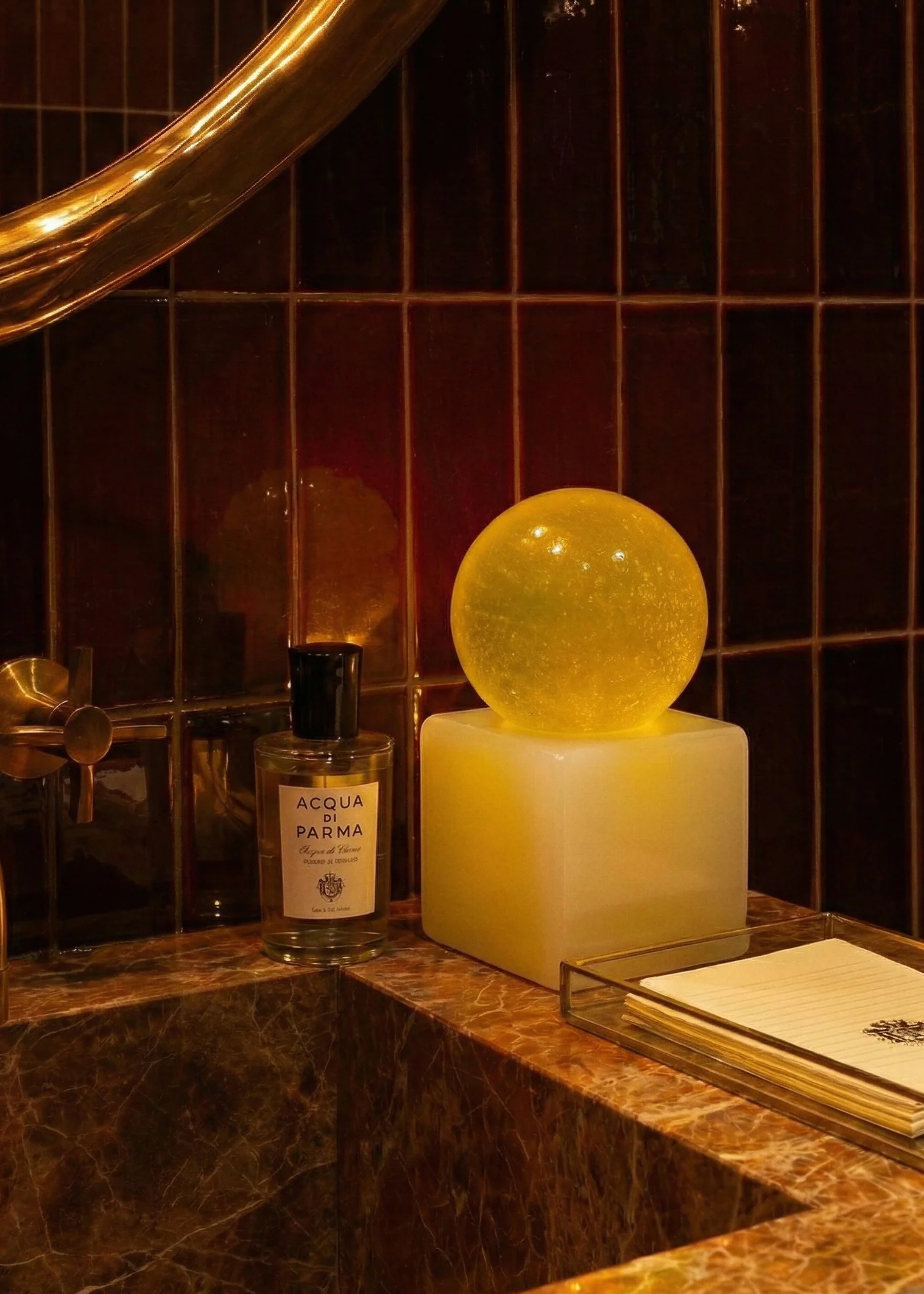 A spherical yellow hand soap on a stone bathroom counter beside an Acqua di Parma perfume against a red tile backsplash