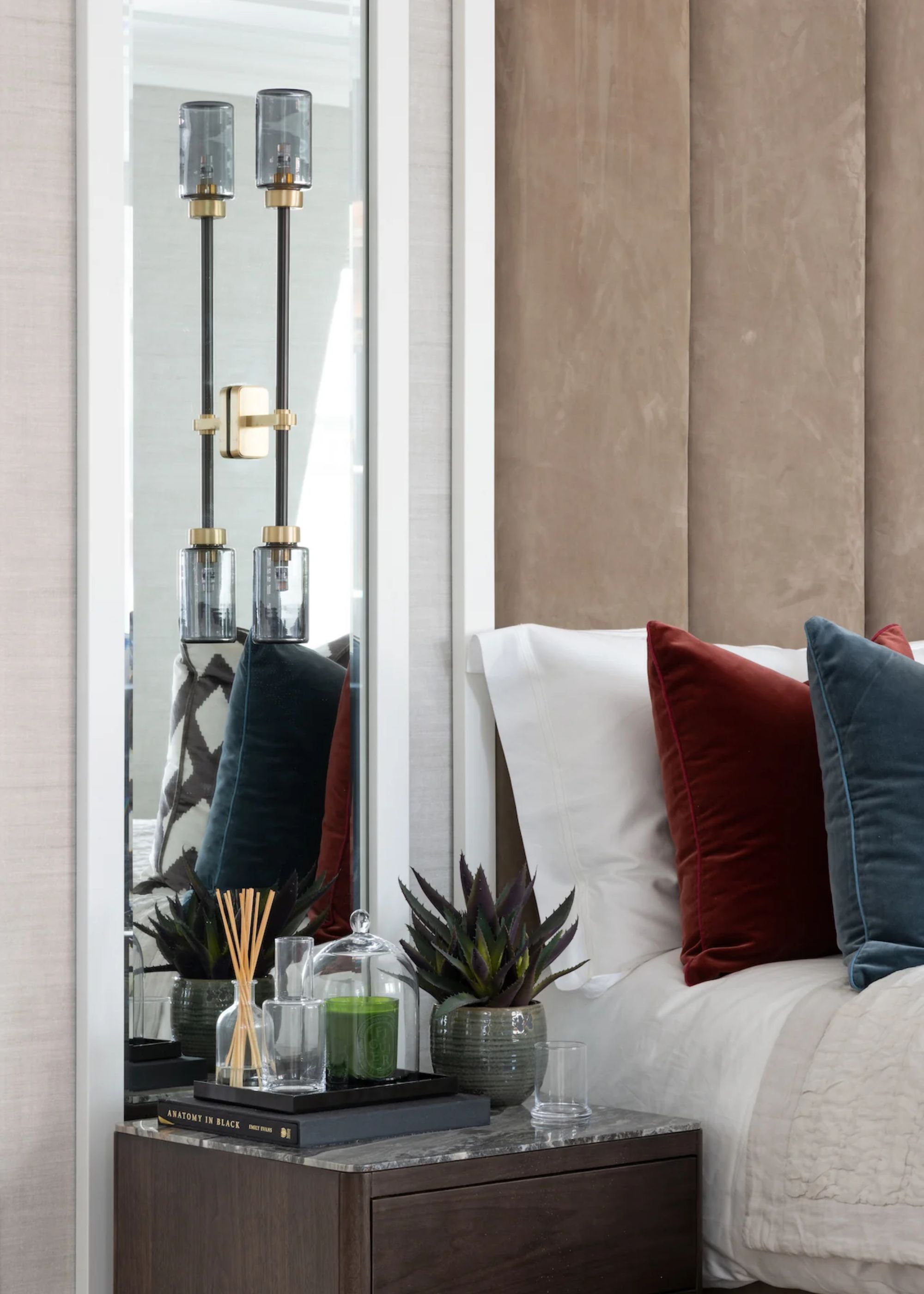 A corner view of a bed with cushions on top and a side table with essentials. Above the side table is a gold and glass wall light