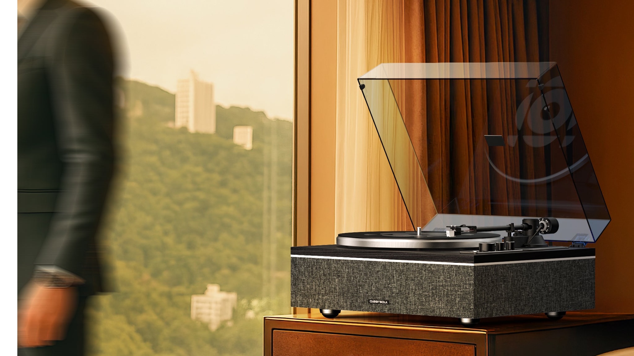 A Qlearsoul ONE-S turntable on a wooden unit in front of a window. A blurred person is moving away on the left of the image.