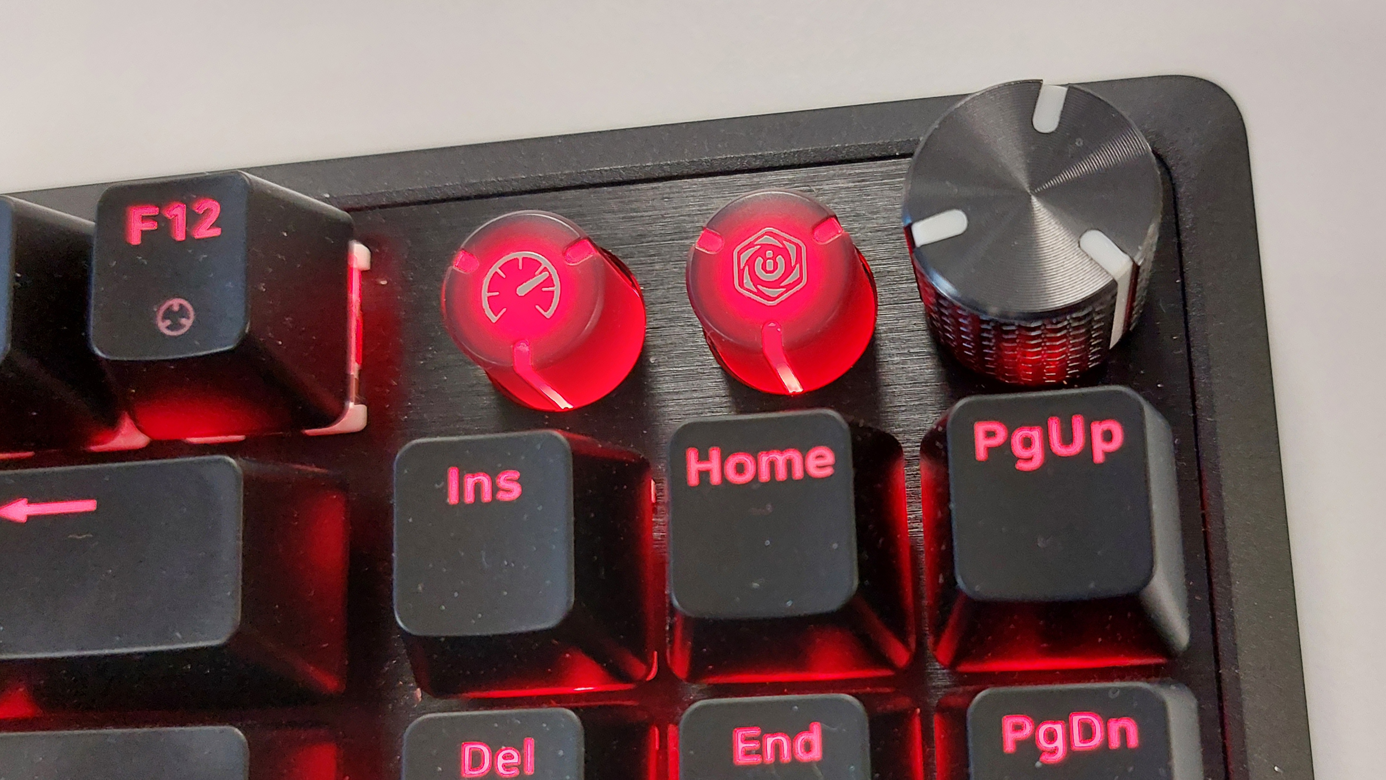 A close up of the Corsair K70 Pro TKL gaming keyboard's two media buttons and rotary control dial. The surroundind keys and two media buttons are lit up in red RGB lighting. The rotary dial is silver metal and stands taller than every surrounding key.