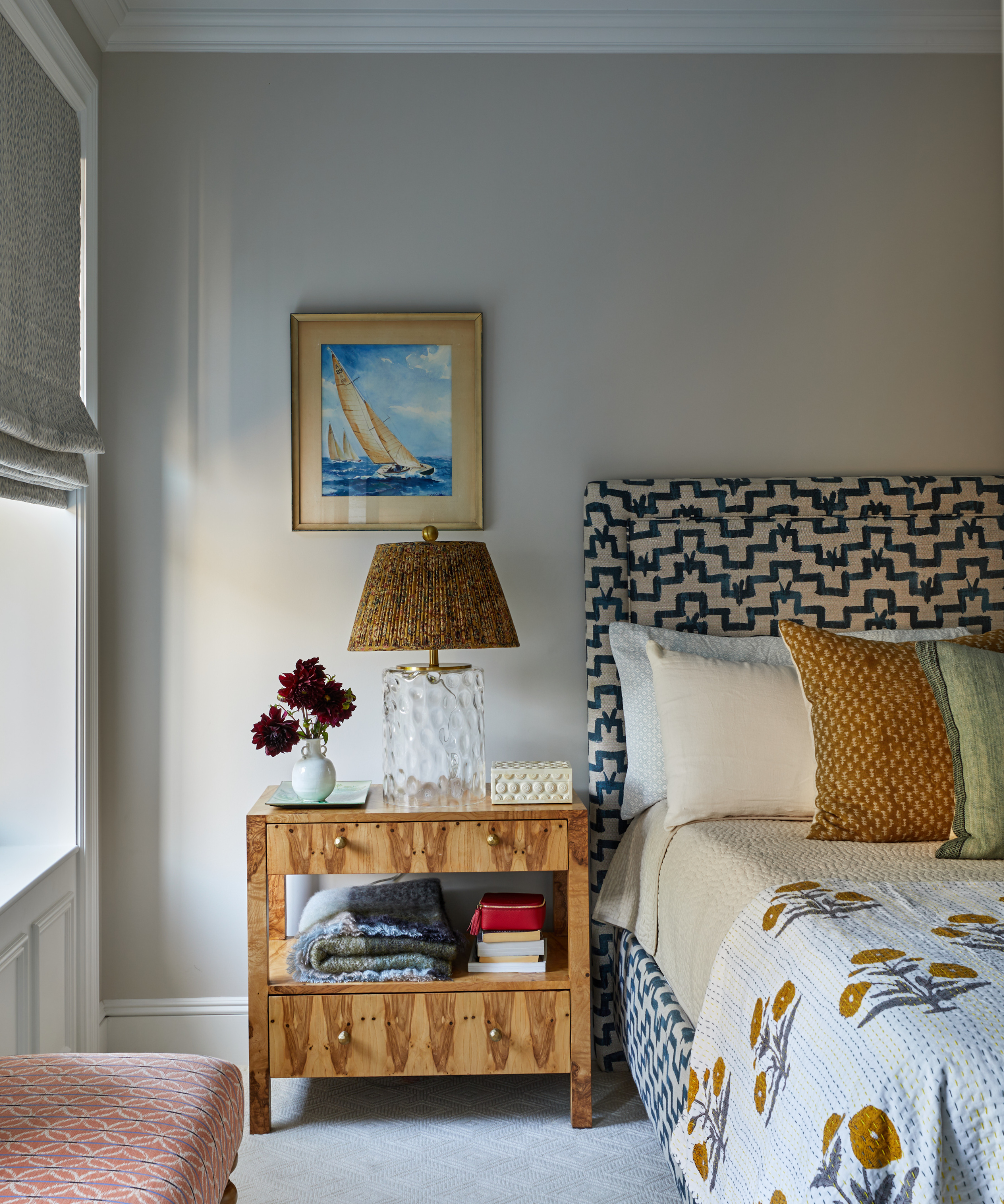 A greige bedroom with a patterned headboard, a wooden side table, and a framed painting of a sailboat
