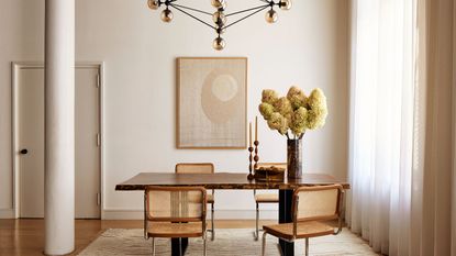Modern dining area with abstract, golden light fixture above a wooden table with four chairs and a potted plant, in neutral room with floor-to-ceiling curtains and a white pillar.