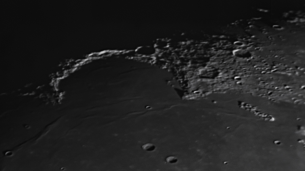 A dark lunar plain is seen bordered by a sickle-like mountain range, which is catching the sun's light close to the terminator separating day from night.