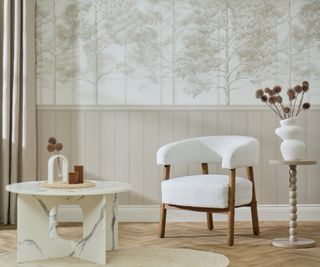 beige and white wallpaper with wood panelled effect on lower half and tree design on top in living room with light wooden floor, cream boucle chair and round white and grey marble coffee table on cream rug