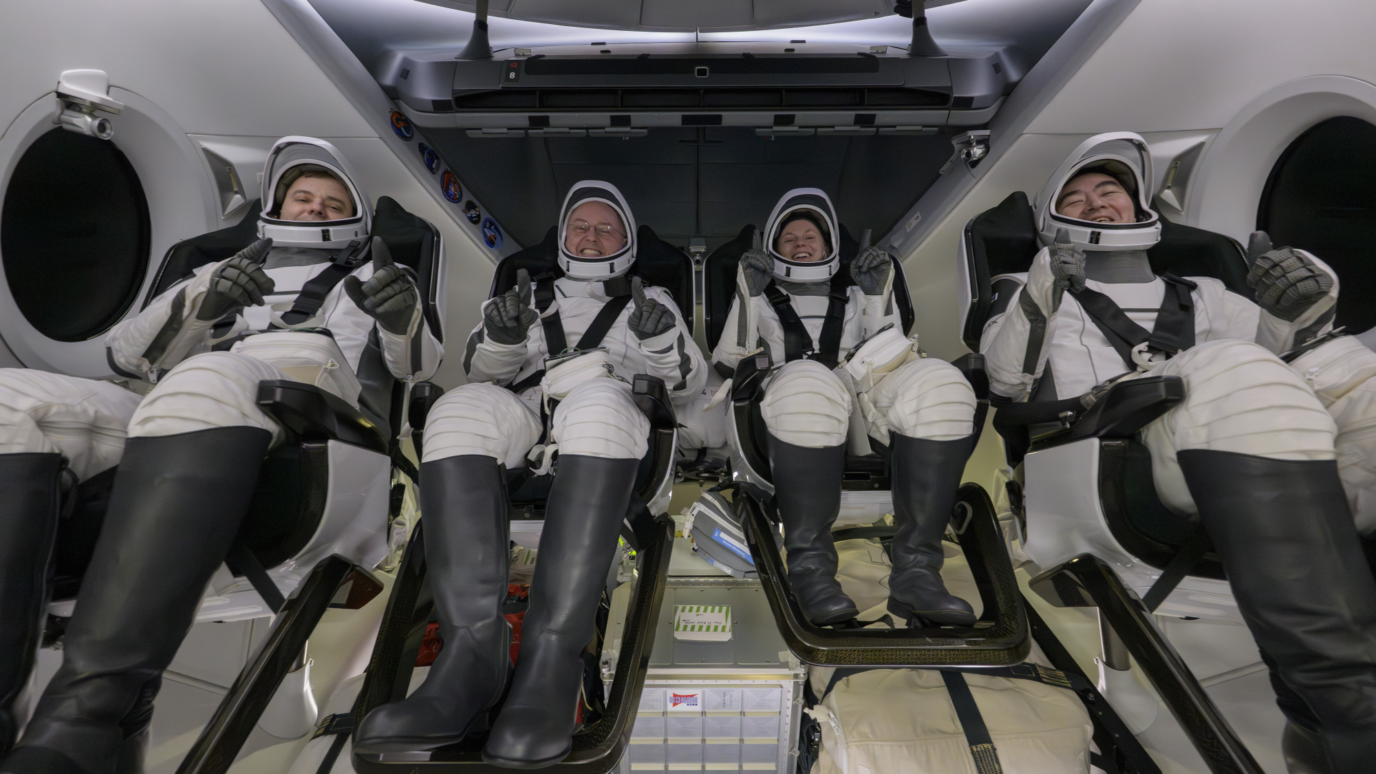 four people in white spacesuits give thumbs up while strapped into seats inside a cramped white cockpit