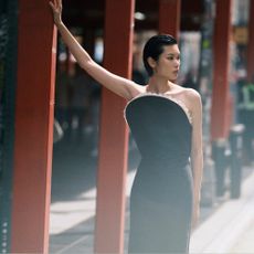 A woman standing on a train station platform wearing a statement black dress from Mytheresa with a hand against a red steel post.