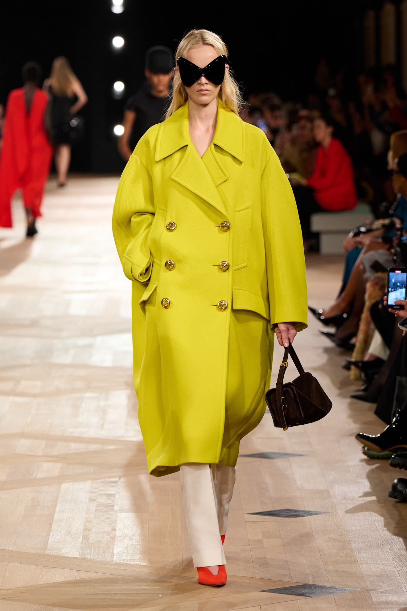 A Balenciaga model wearing an oversize double-breasted yellow coat styled with white slim-fit trousers, red pointed heels, a brown handbag, and black oversize bug-eye sunglasses.