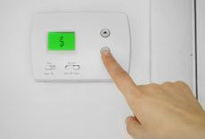 Finger pressing a button on a thermostat that displays a dollar sign, indicating rising energy costs.