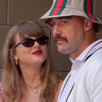 Taylor Swift, wearing sunglasses, smiles at Travis Kelce, who is wearing a Gucci bucket hat and white polo shirt