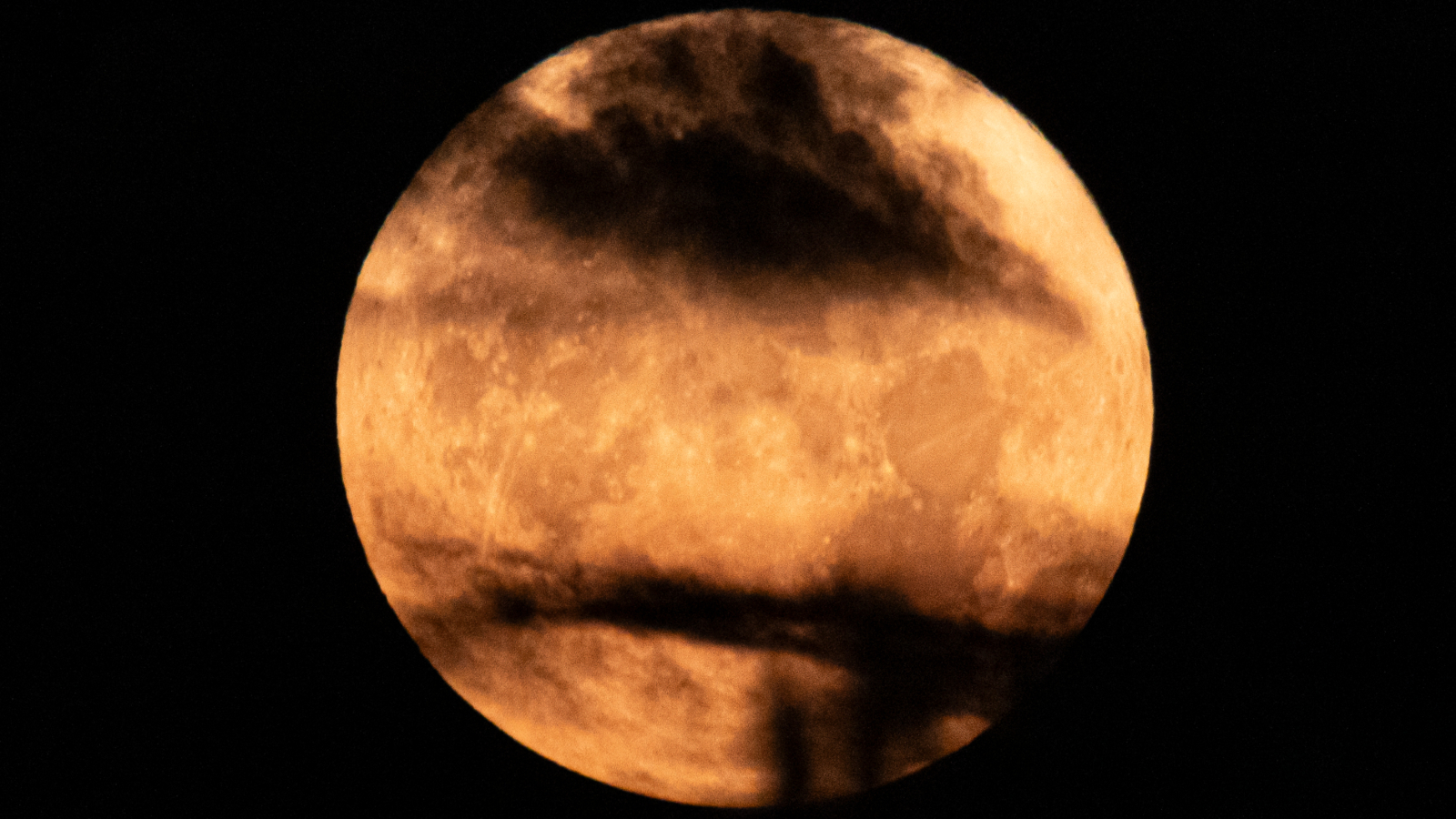 A yellow-orange full moon is pictured against a black night sky. Dark clouds obscure parts of the lunar disk and dark patches called ‘lunar maria’ can be seen covering swathes of the surface.