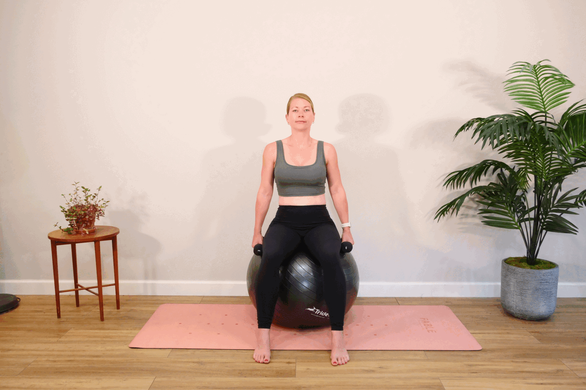 A woman sits on an exercise ball holding dumbbells in her hands and lifts and raises her arms to level with her shoudlers