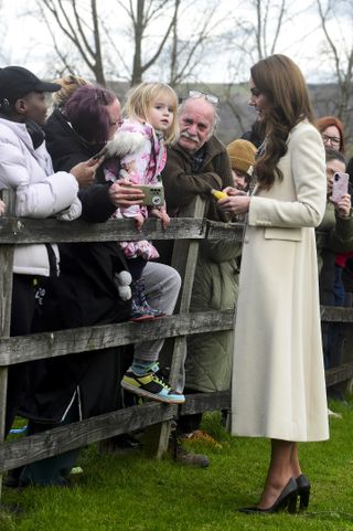 Kate Middleton wears a long cream coat with a dogtooth print dress and speaks to a young girl in the crowd