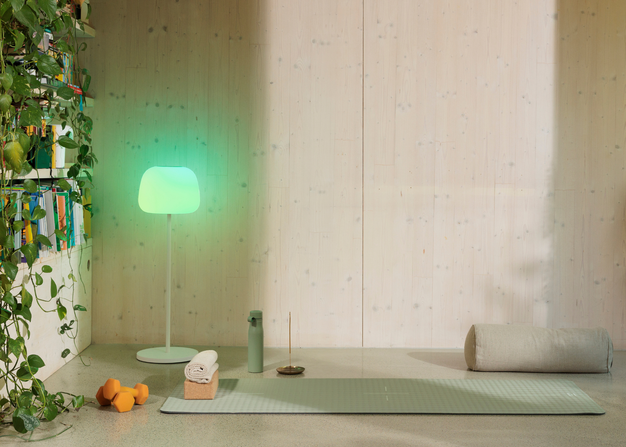 A wellness room with a floor lamp emitting green light beside a yoga mat and brick, an incense holder, orange weights, and a bolster pillow