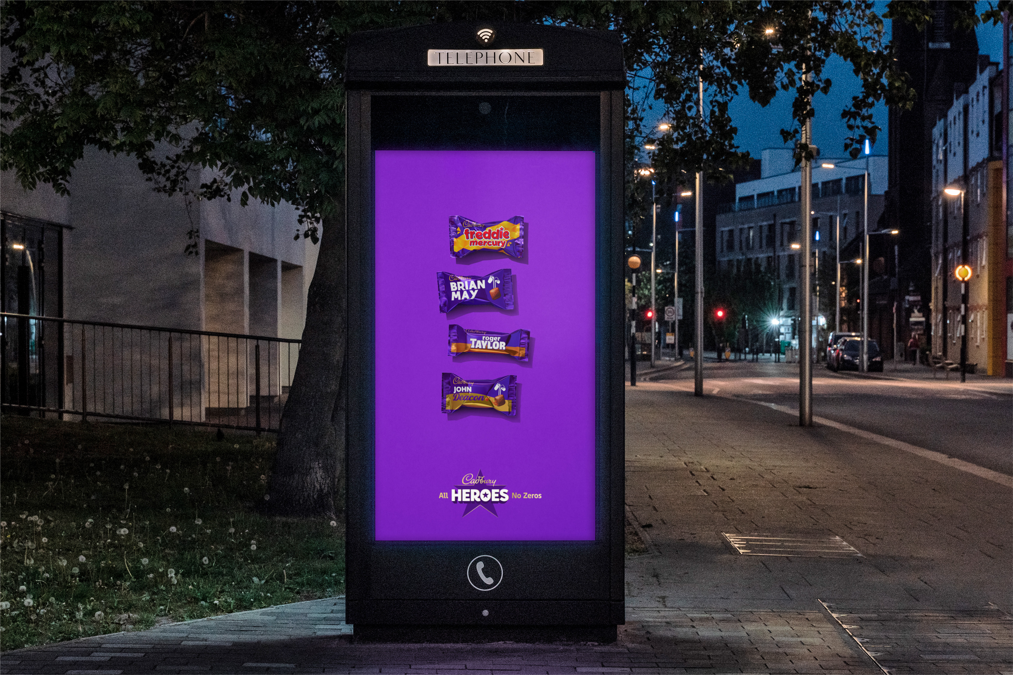 An outdoor digital advertisement on a telephone box at night featuring four miniature Cadbury&#039;s chocolate bars named after the band members of Queen (Freddie Mercury, Brian May, Roger Taylor, John Deacon) stacked vertically over a purple background with the slogan &amp;quot;Cadbury All HEROES No Zeros.&amp;quot;