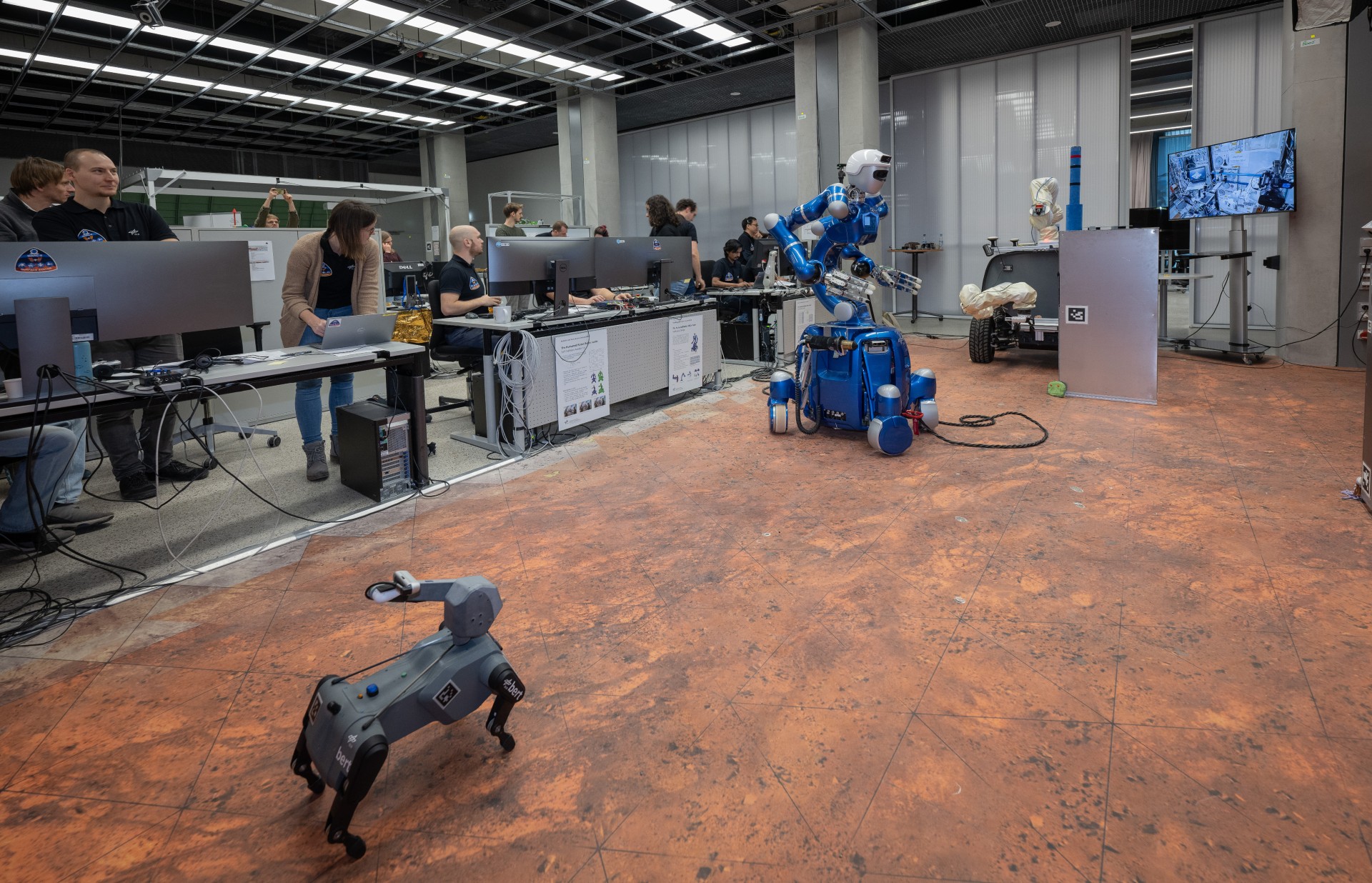 a dog-like robot and a humanoid robot on wheels roll around a laboratory surrounded by scientists in t-shirts standing at computers