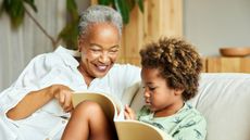 An older woman sits on the sofa with her grandson as they read a book together.