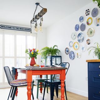 White dining room with orange table, black chairs and collection of decorated plates on wall