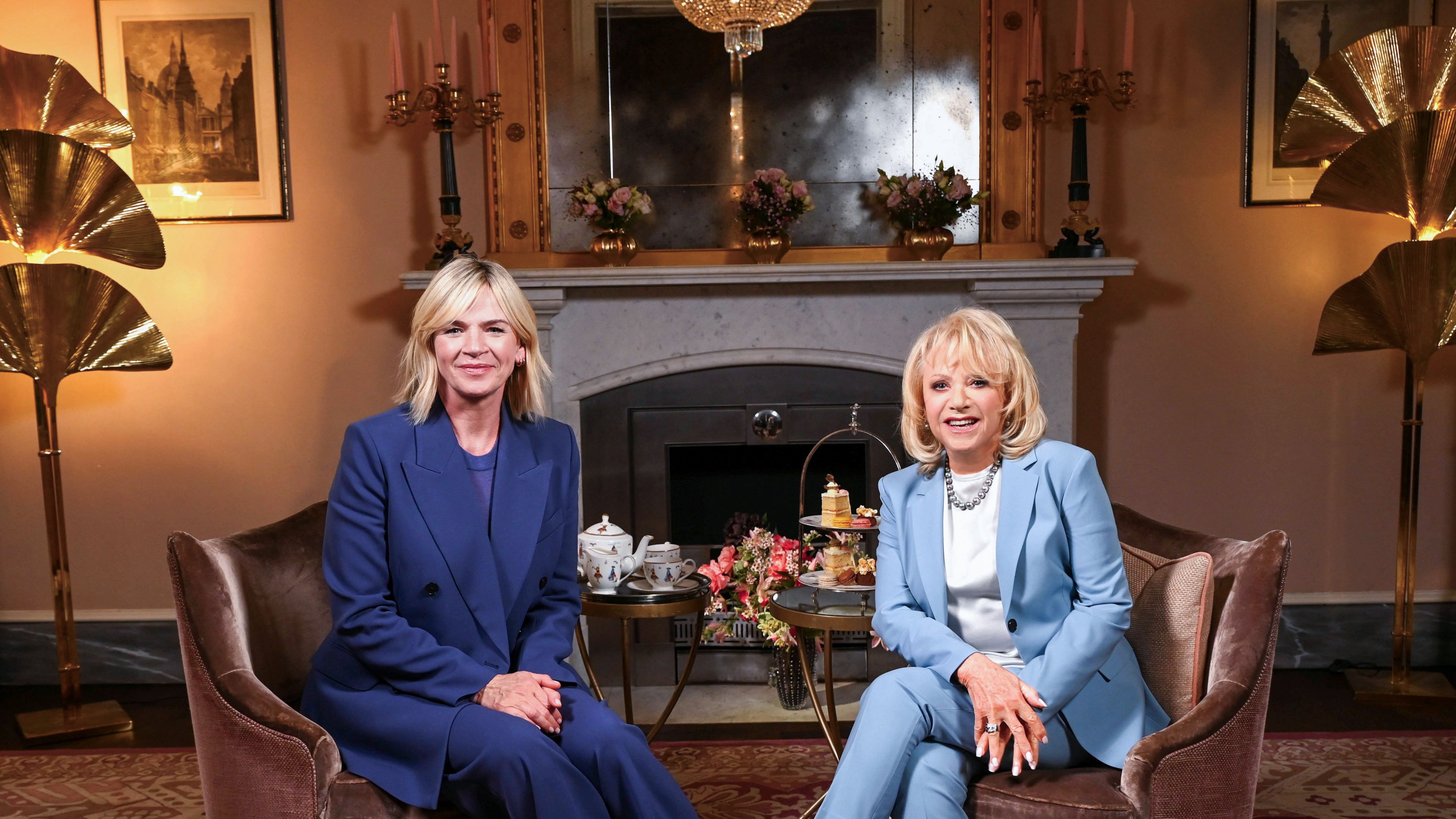 Zoe Ball and Elaine Paige both in blue suits sit by a fireplace with cakes on a table in Elaine Paige: 60 Years on Stage