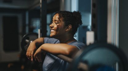 Smiling woman in workout clothes in gym, leaning on a barbell