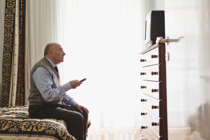 A man sits on a bed while watching television. 