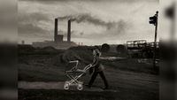 Black and white photograph of a woman pushing a pram along a bleak industrial road, with factory chimneys smoking in the background.